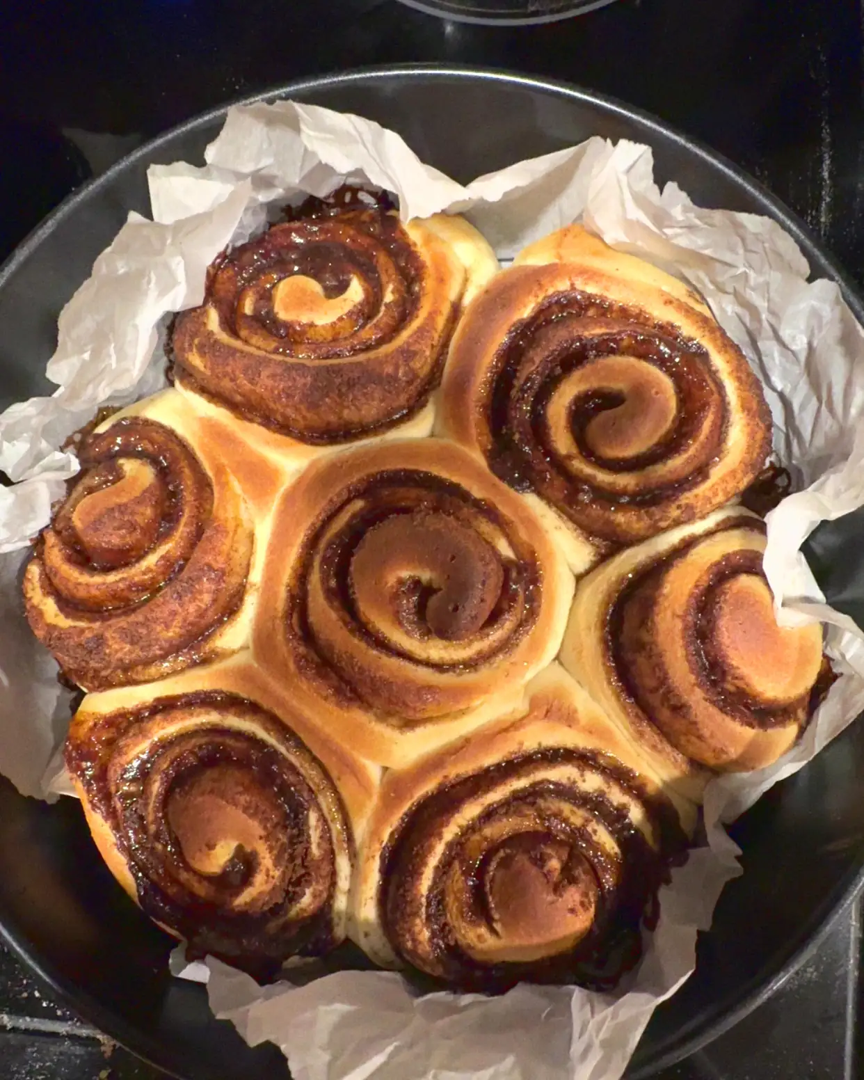Overhead view of a round pan of unfrosted cinnamon rolls, seven spirals packed together on crumpled parchment, on a black stovetop.