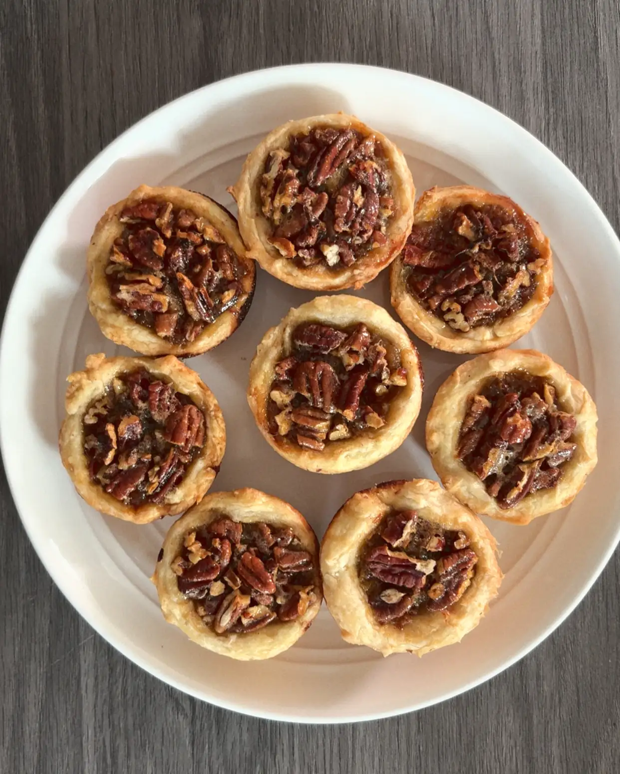 Eight small pecan tartlets, seven in a ring and one in the center, on a white plate on a wood-grain table.