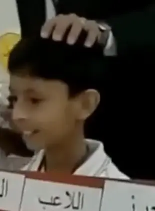 Mohammed as a boy, smiling, the teacher’s hand resting on his head and the top of his red board of word cards in front of him, in a lesson at Qurtubah Schools, Jeddah.