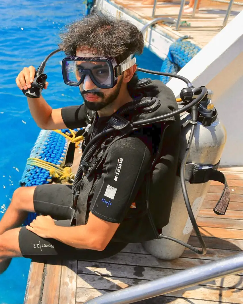 Mohammed in scuba gear on the wooden stern of a dive boat, mask on and regulator raised in one hand, looking up at the camera.